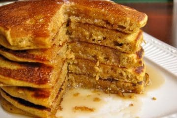 A stack of Pumpkin Pancakes featuring five pancakes, and a cutaway view inside the stack, drizzled with maple syrup.