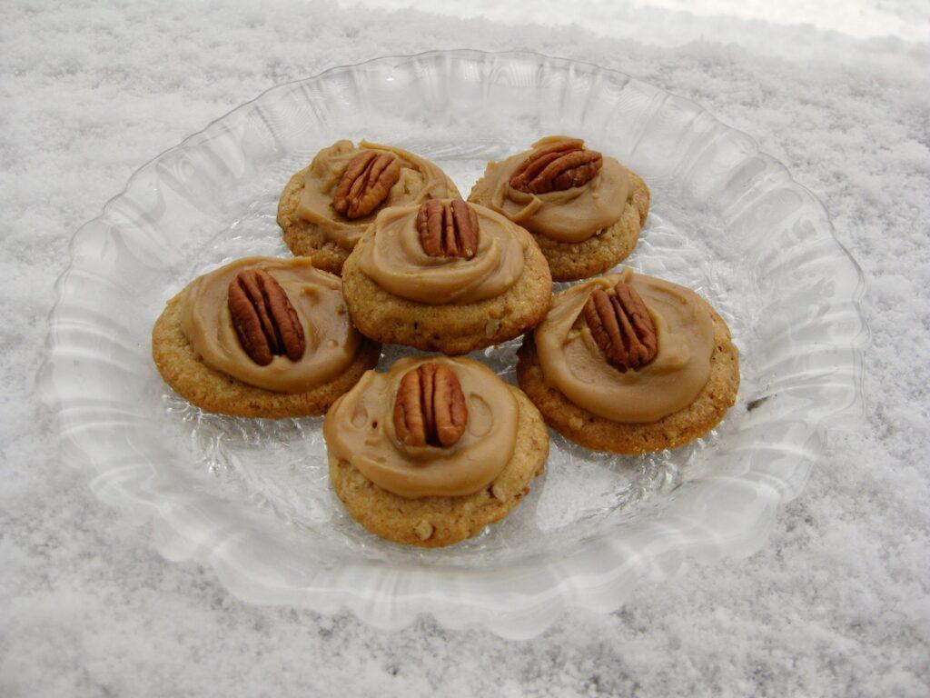 Pecan Praline Cookies with Brown Sugar Frosting, topped with pecan halves, are resting on a snowy ledge.