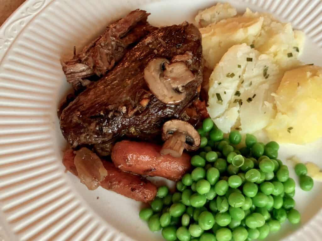 Beef Pot Roast served on a plate beside green peas and herbed potatoes.