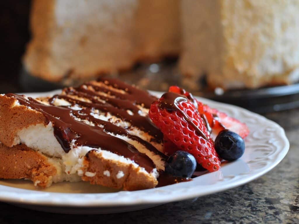 A thin slice of Angel Food Cake topped with fresh berries and chocolate ganache in front of the sliced Angel Food Cake.