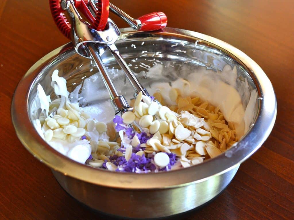 Batter for Lavender Meringue Cookies in a bowl with fresh lavender petals, white chocolate morsels and toasted almonds, mixed with a rotary egg beater.