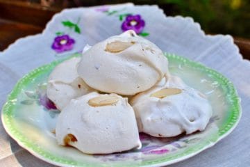 Lavender Meringues with White Chocolate and Almonds stacked on an antique plate beside an embroidered napkin.
