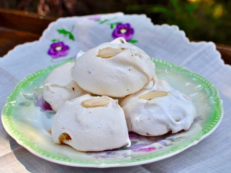 Lavender Meringues with White Chocolate and Almonds stacked on an antique plate beside an embroidered napkin.