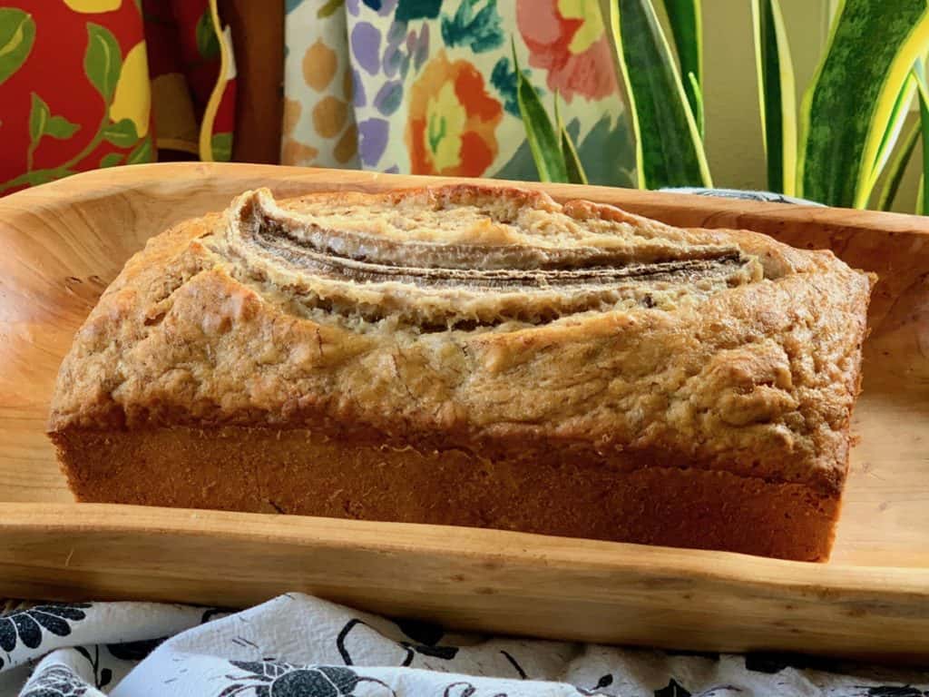An 8-inch loaf of Banana Bread on a wooden tray in front of aprons and leaves.