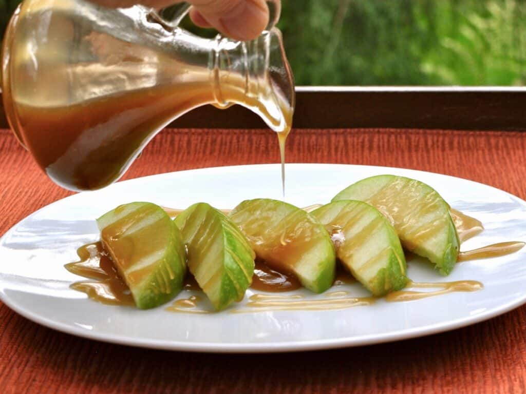 Caramel Sauce in a small cruet is being drizzled over a plate of sliced apple wedges.