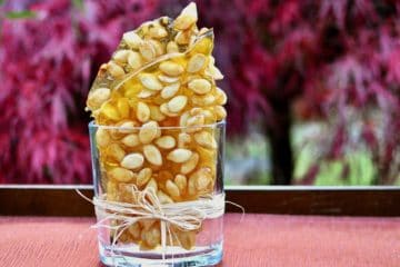Pieces of Pumpkin Seed Brittle arranged in a clear glass container in front of autumn leaves.