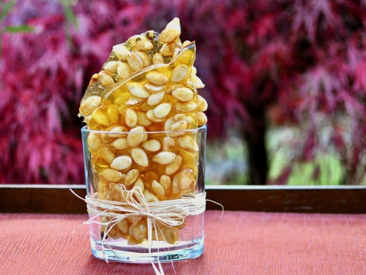 Pieces of Pumpkin Seed Brittle arranged in a clear glass container in front of autumn leaves.