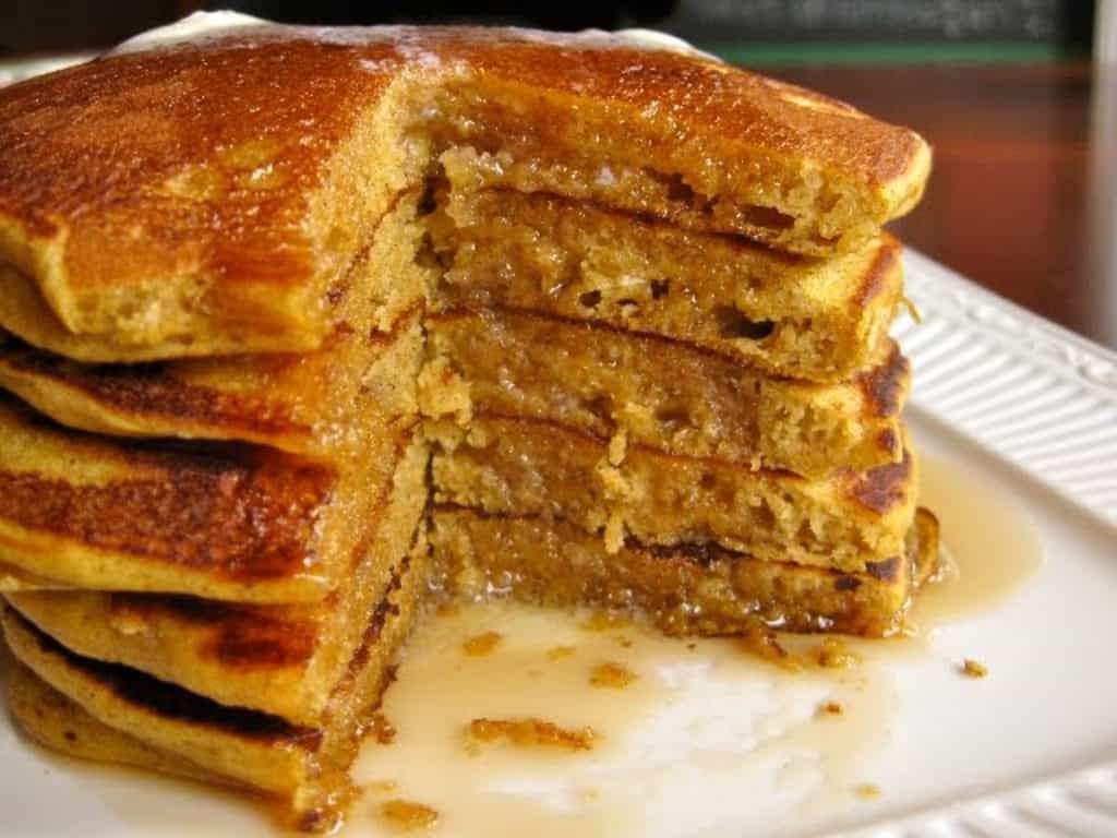 A tall stack of pumpkin pancakes with a wedge cut away, drenched in maple syrup.