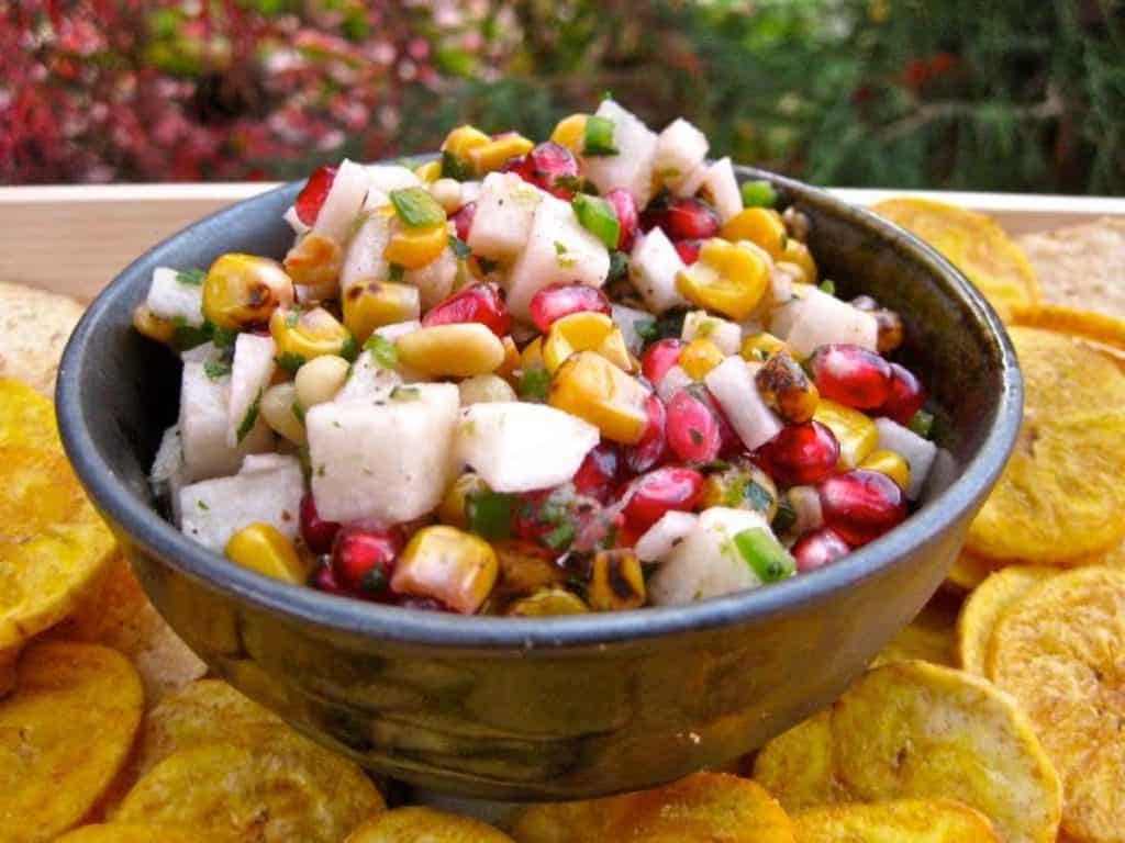 Pomegranate and Roasted Corn Salas in a small bowl surrounded by Plaintain Chips.  A fantastic starter for your Thanksgiving menu. 