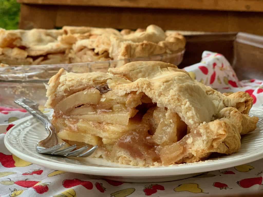 A slice of Rustic Apple Pie on a plate beside a fork with the cut pie in the background