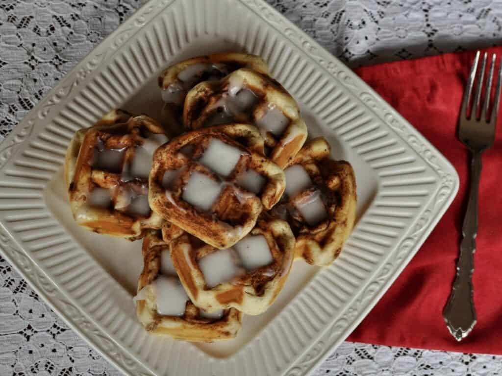 Cinnamon Roll Waffles stacked on a square plate and drizzled with icing, set on a lace cloth with a red napkin.