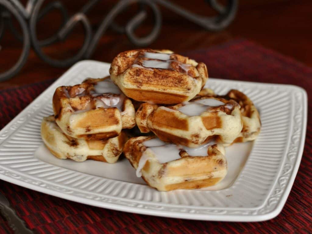Sweet and Sassy Cinnamon Roll Waffles stacked on a square plate and drizzled with icing. 