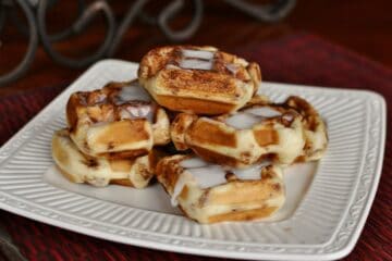Sweet and Sassy Cinnamon Roll Waffles stacked on a square plate and drizzled with icing.