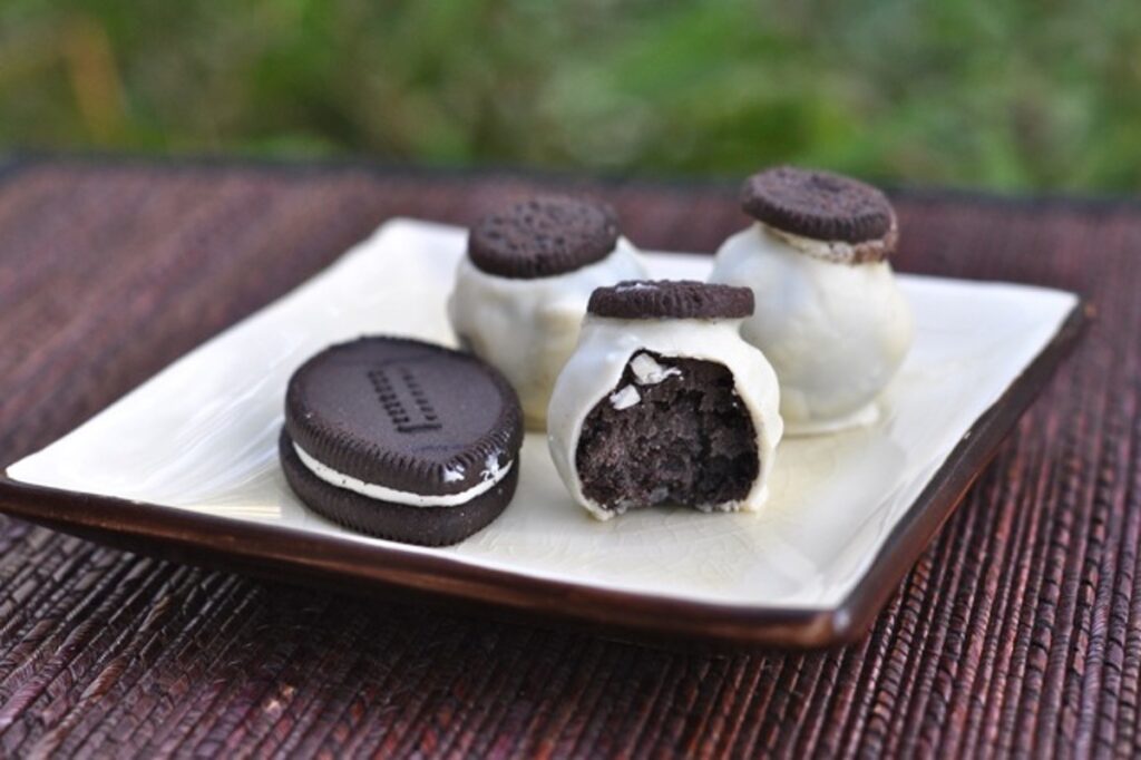 A football shaped Oreo Cookie and several mini Oreo topped bite-sized Oreo Truffles are displayed on a small ceramic plate. One Oreo Truffle shows the Oreo Cream Cheese filling.