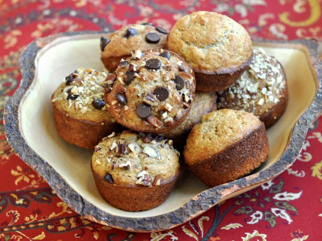 A variety of Banana Muffins, some topped with nuts, others with chocolate chips and some plain, served in a uniquely shaped rustic wooden bowl.