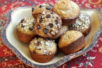 A variety of Banana Muffins, some topped with nuts, others with chocolate chips and some plain, served in a uniquely shaped rustic wooden bowl.