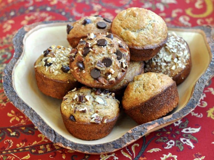 A variety of Banana Muffins, some topped with nuts, others with chocolate chips and some plain, served in a uniquely shaped rustic wooden bowl.