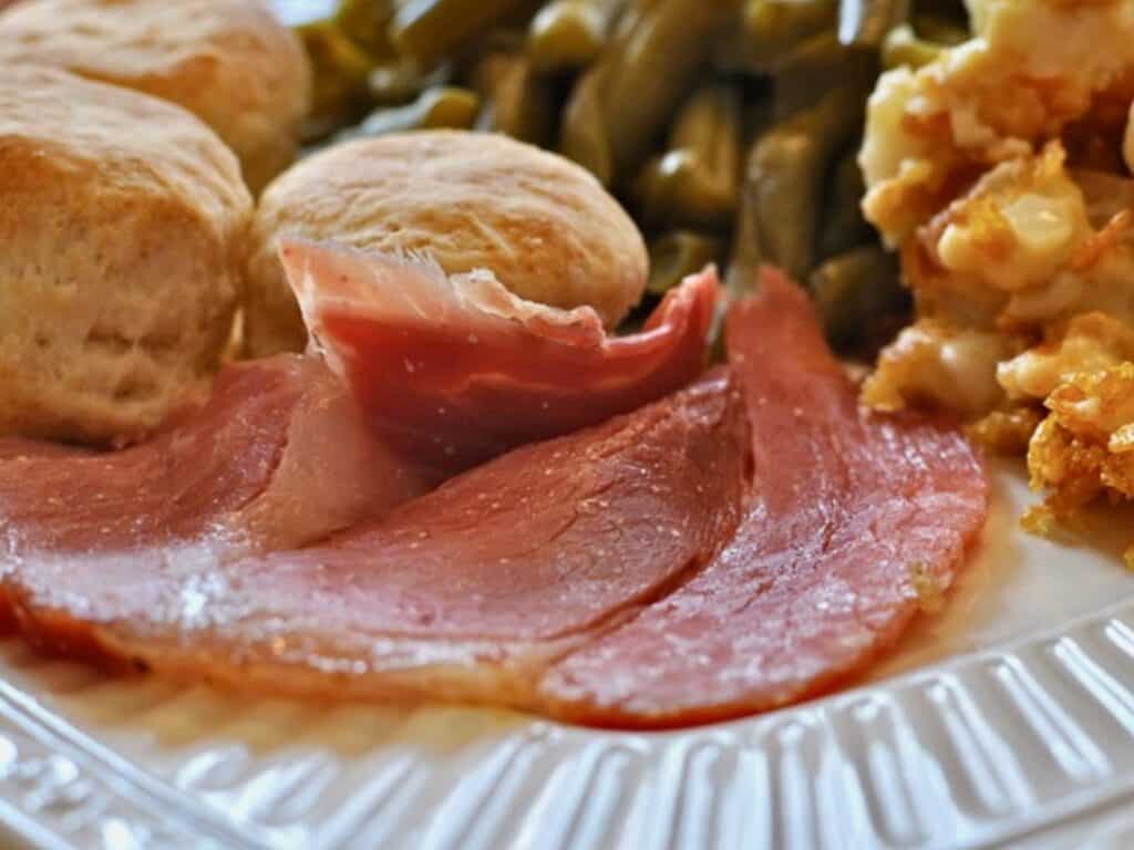 A slice of Kentucky Country Ham is served on a dinner plate with Biscuits, Southern Style Green Beans and Cornflake Casserole.