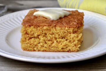 A slice of Back to Basics Cornbread topped with a melting pat of butter and served on a white plate.