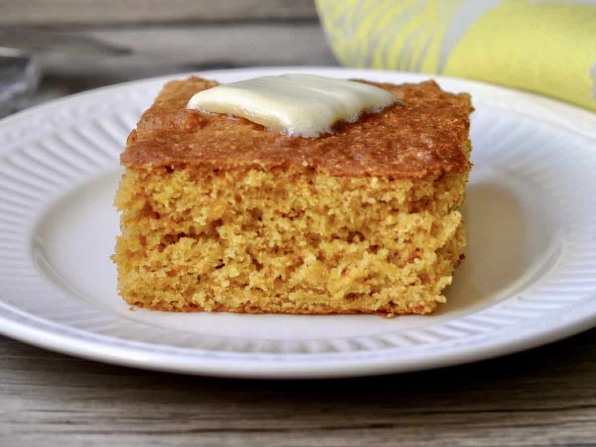 A slice of Back to Basics Cornbread topped with a melting pat of butter and served on a white plate.