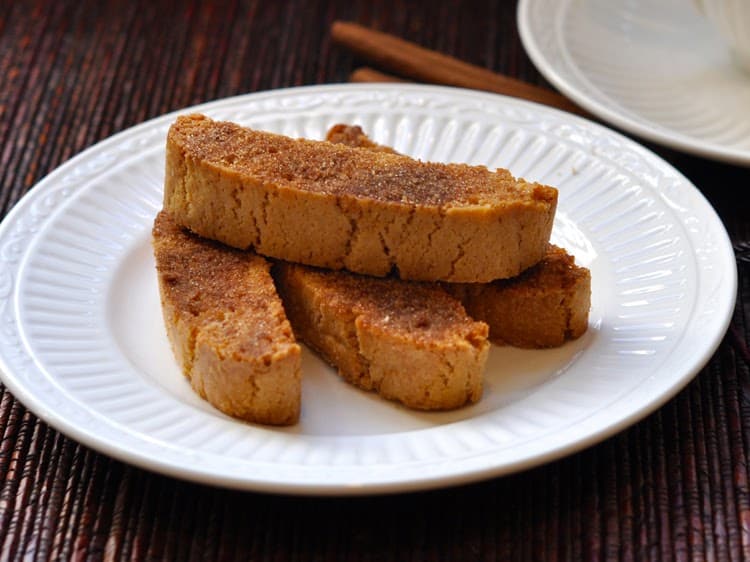 Four Cinnamon Toast Biscotti arranged on a white saucer beside cinnamon sticks and a coffee cup.