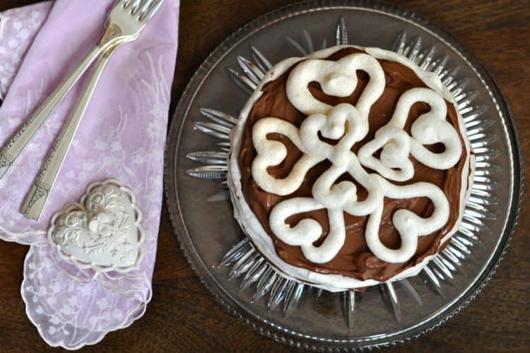 Meringue Torte with Chocolate filling on a glass plate beside an embroidered napkin, a heart shaped ornament and two silver forks.