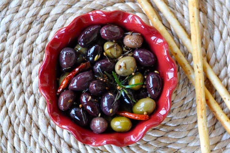 Herb Spiced Olives with, rosemary, coriander and fennel seeds, served in a small red dish with breadsticks on the side.