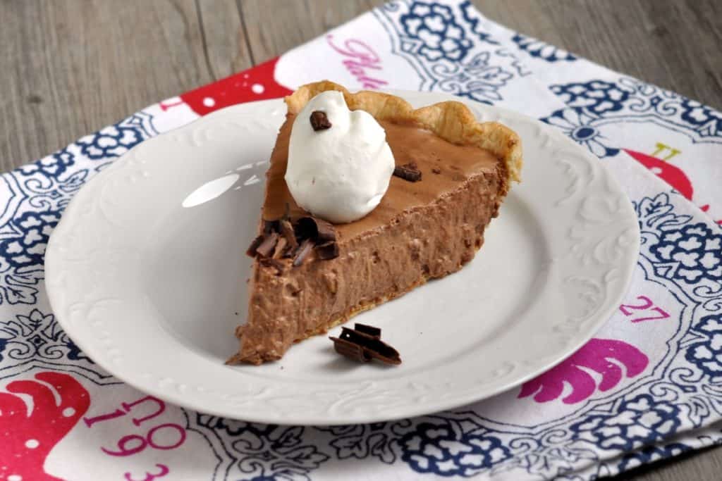 A slice of Aunt Hen's Chocolate Bar Pie topped with whipped cream and chocolate flecks on a white dessert plate.