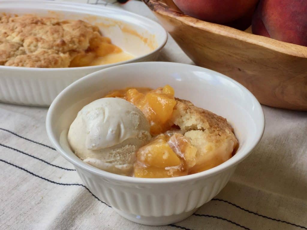 Peach Cobbler for Two served in a bowl and topped with a scoop of vanilla ice cream beside a bowl of ripe peaches.