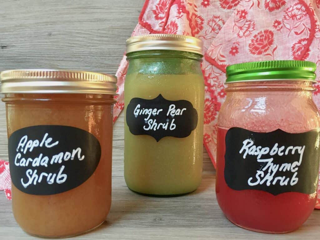 A variety of shrubs, including Pear Ginger Shrub, in mason jars with blackboard labels displayed in front of a vintage apron.