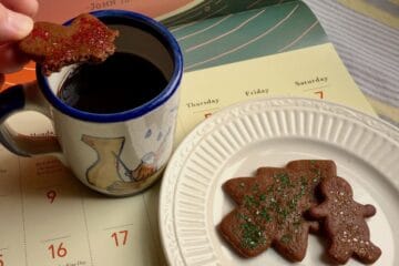 A plate of Gingerbread Cookies topped with sparkling sugar sprinkles are resting on a calendar page. One has a bite taken from it and is being dipped in a cup of coffee.