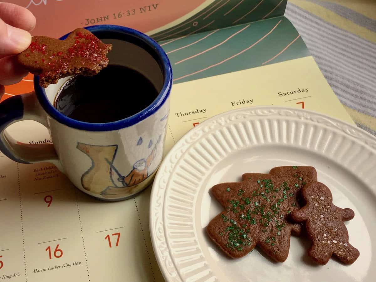 A plate of Gingerbread Cookies topped with sparkling sugar sprinkles are resting on a calendar page. One has a bite taken from it and is being dipped in a cup of coffee.