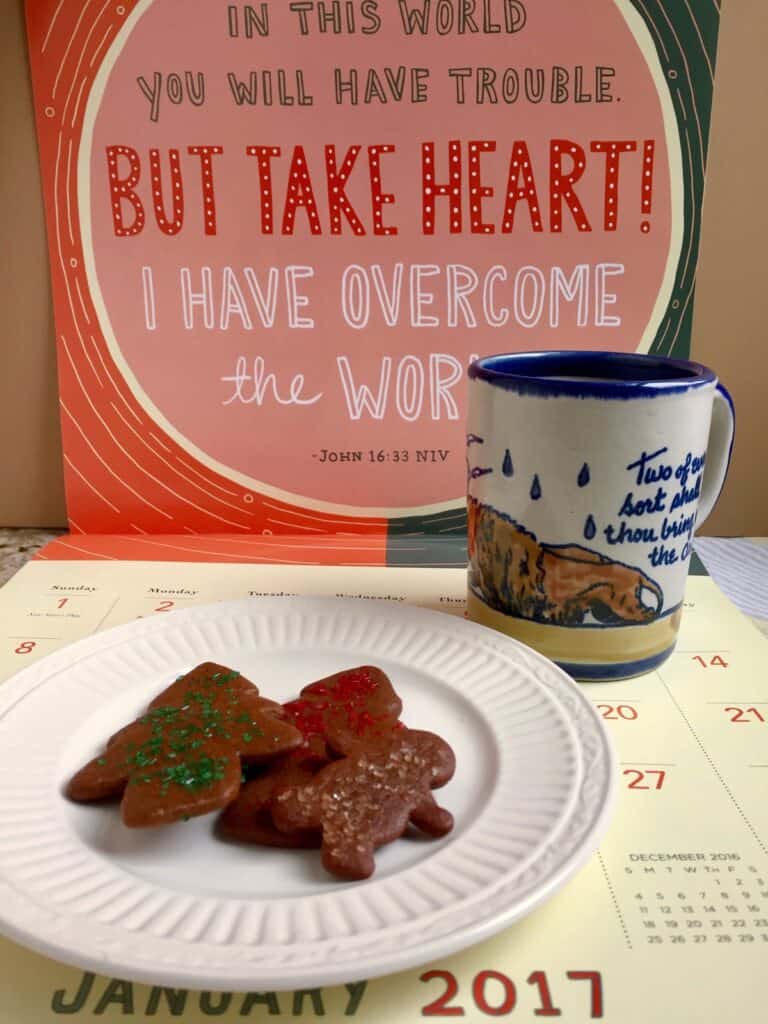 A plate of Gingerbread Cookies topped with sparkling sugar sprinkles are resting on a calendar page with a mug of coffee. A Bible verse on the calendar reads, "In this world you will have trouble but take heart! I have overcome the world." - John 16:33 NIV.