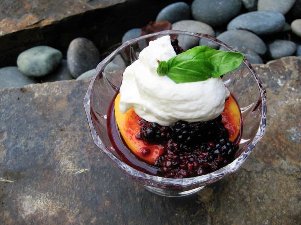 Summer Berry Basil Sauce made with fresh blackberries over a ripe peach half topped with whipped cream and a sprig of basil in a glass dish placed on a rocky ledge
