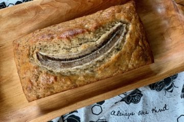 A loaf of Banana Bread, topped with a split banana, on a wooden tray. The tea towel beneath it says, "Always bee kind!"