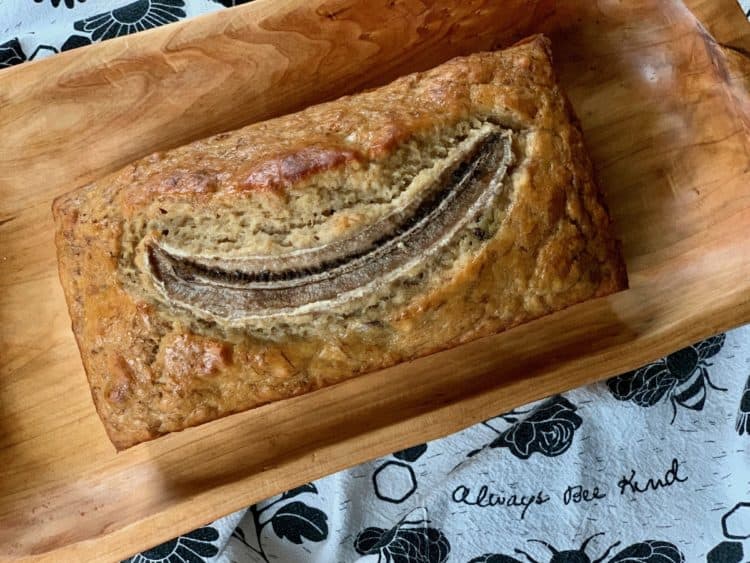 A loaf of Banana Bread, topped with a split banana, on a wooden tray. The tea towel beneath it says, "Always bee kind!"