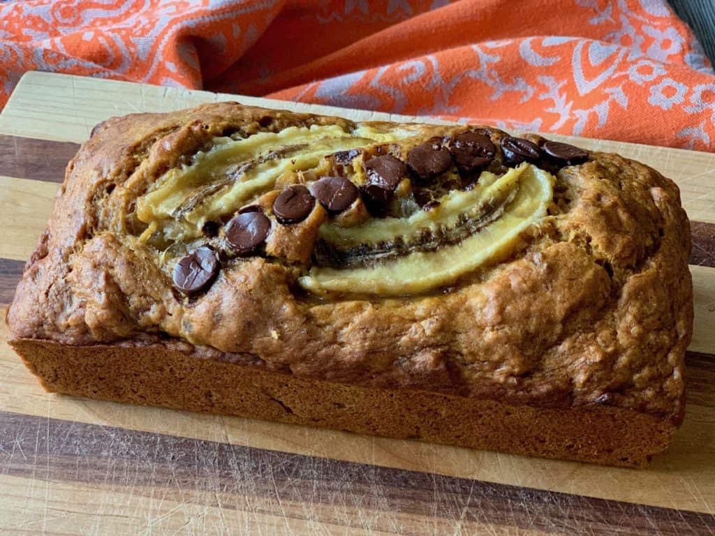 A loaf of Banana Pumpkin Bread, topped with a split banana and chocolate chips, cooling on a striped wood cutting board.