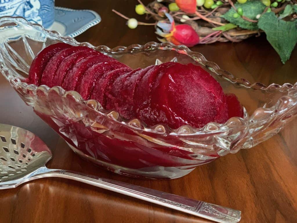 Smooth Cranberry Sauce frozen and sliced in an Anchor Hocking pressed glass serving boat beside a pierced silver serving spoon.