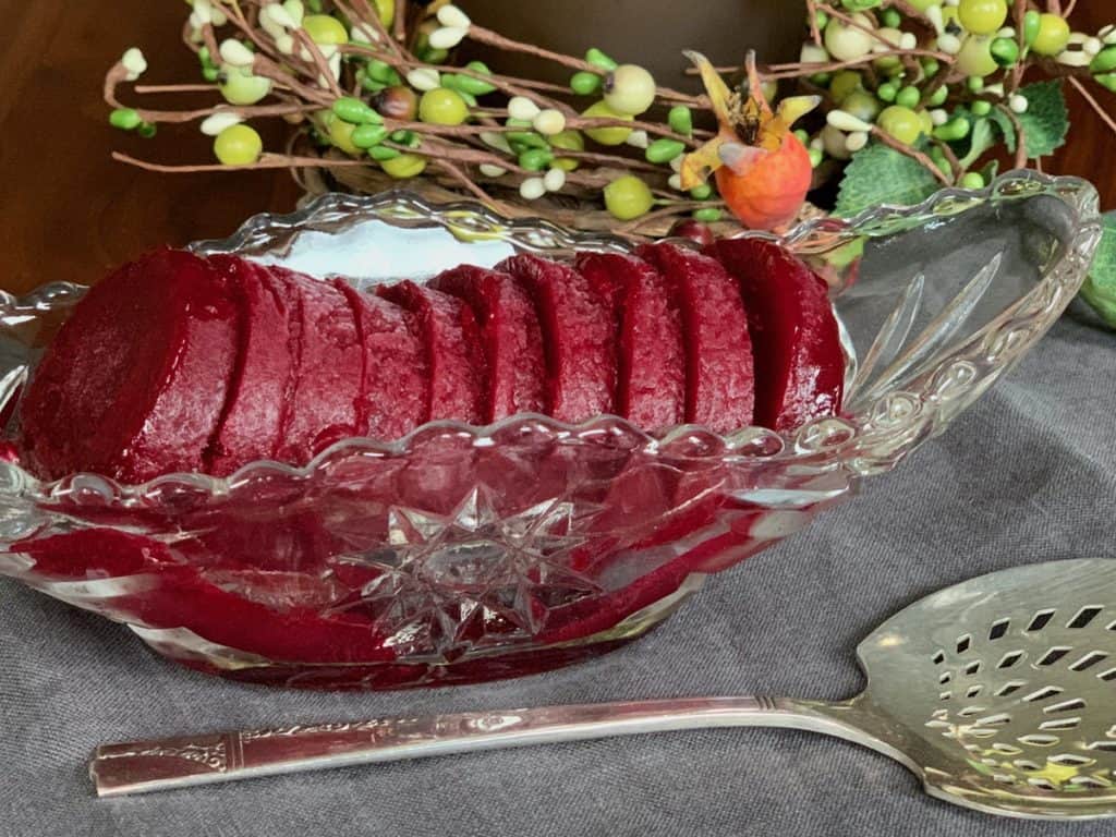Smooth Cranberry Sauce frozen and sliced in an Anchor Hocking pressed glass serving boat beside a pierced silver serving spoon. 