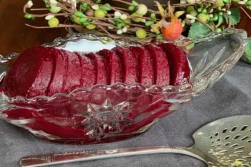 Smooth Cranberry Sauce frozen and sliced in an Anchor Hocking pressed glass serving boat beside a pierced silver serving spoon.