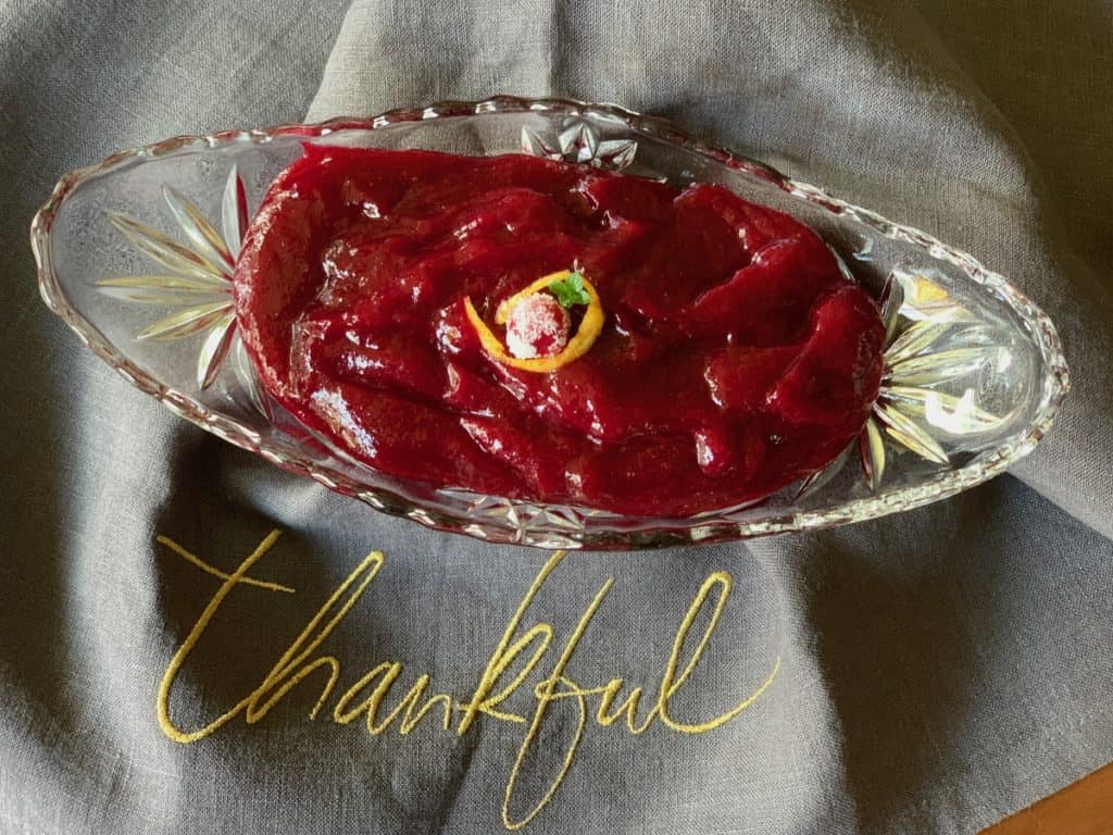 Smooth Cranberry Sauce, garnished with a sugared cranberry, in a pressed glass serving boat on a cloth embroidered with the word "thankful".