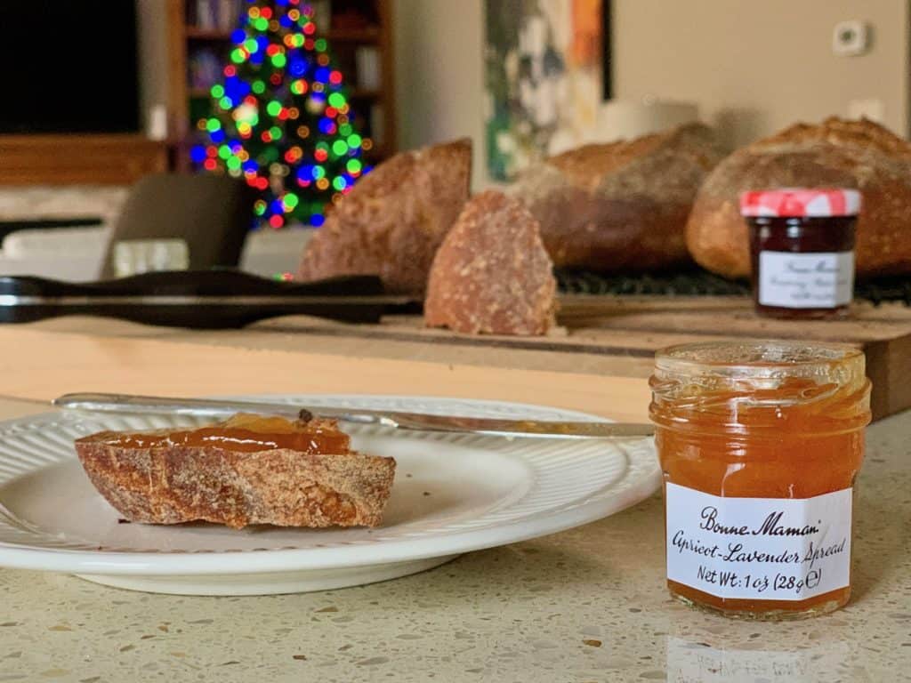 A jar of Apricot Lavender Fruit Spread open and spread on a slice of toasted Sourdough Bread with a lit Christmas Tree in the background.