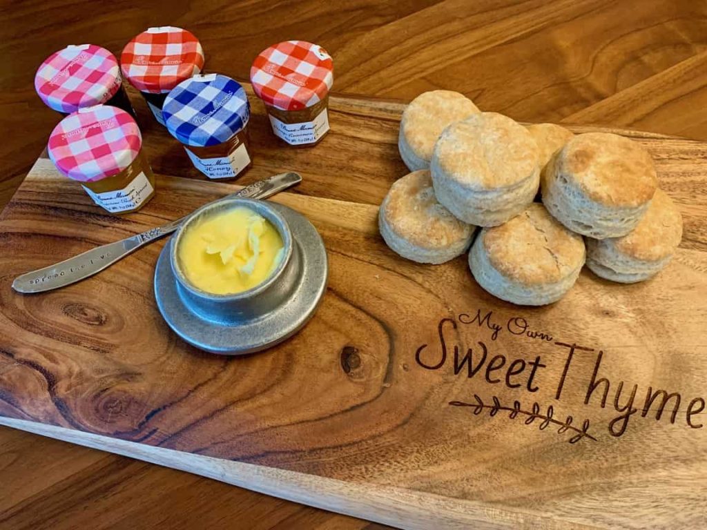 Several small jars of fruit spread and a container of butter beside a stack of Uncle Hal's Biscuits on our My Own Sweet Thyme cutting board.