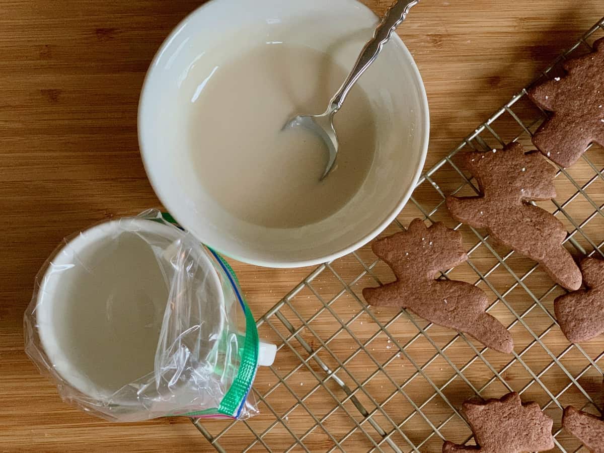 Vanilla Icing mixed in a small bowl beside a teacup fitted with a ziploc bag to use for a piping bag to decorate the ginger spice cookies on the rack by its side.