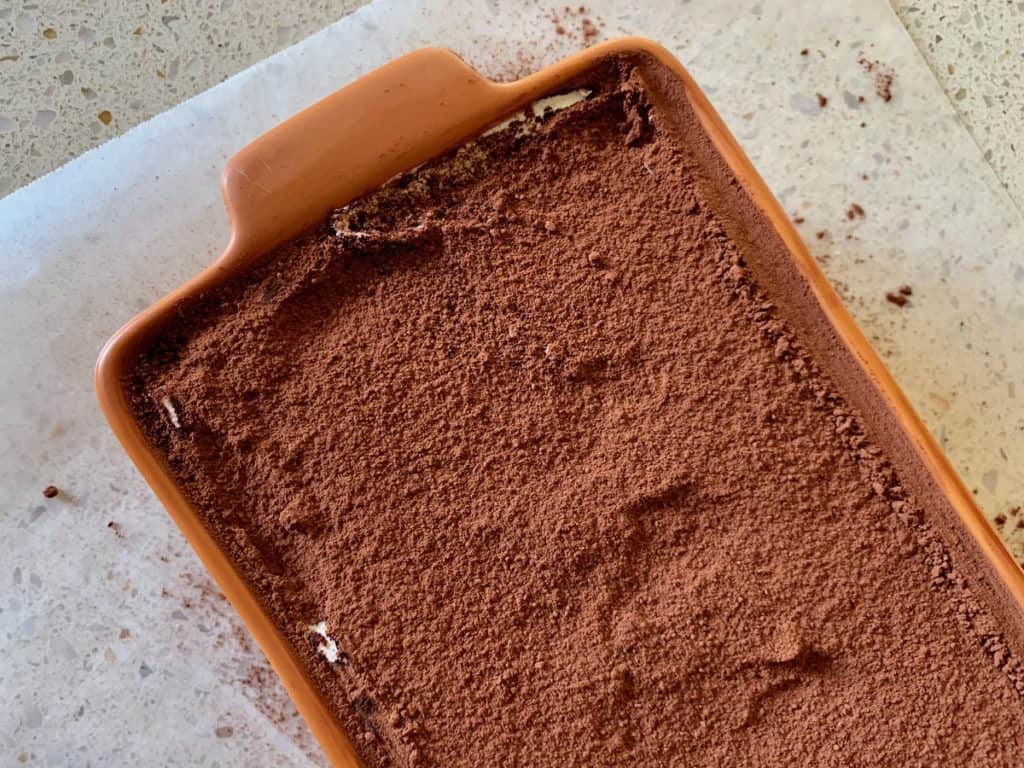 Simple Tiramisu in a ceramic serving dish set on a piece of wax paper to catch any stray bits of cocoa powder while dusting the cake. 
