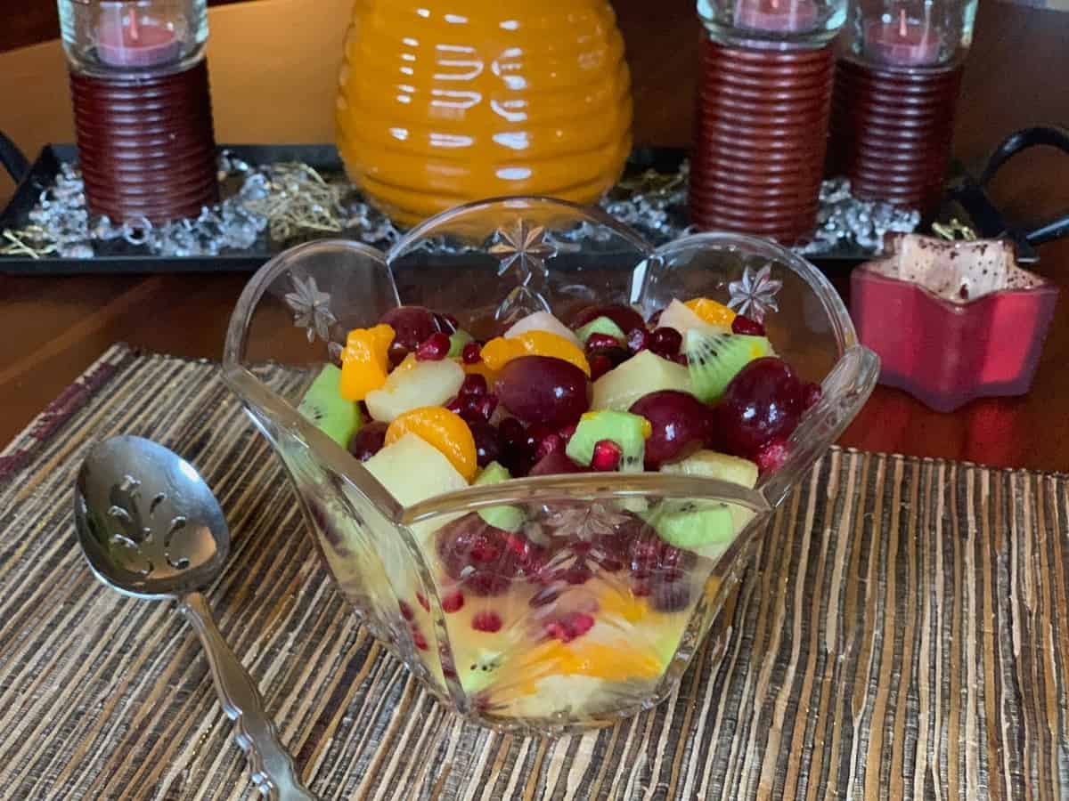 Simply Festive Fruit Salad, served in a pretty glass bowl, with holiday decorations in the background.