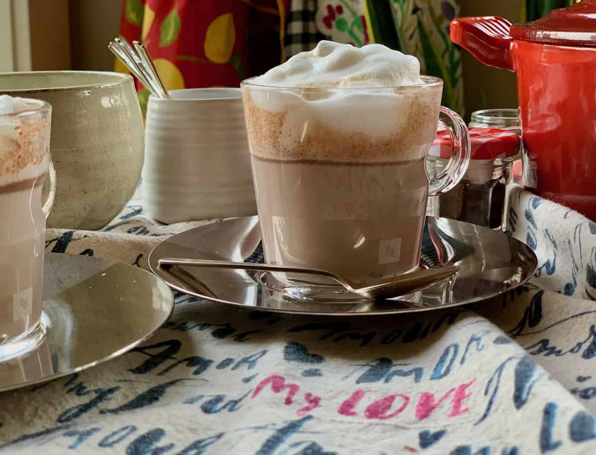 Fudgy Hot Chocolate prepared in a glass cup beside a small Hot Chocolate Kit and several utensils on a "Love" tea towel.
