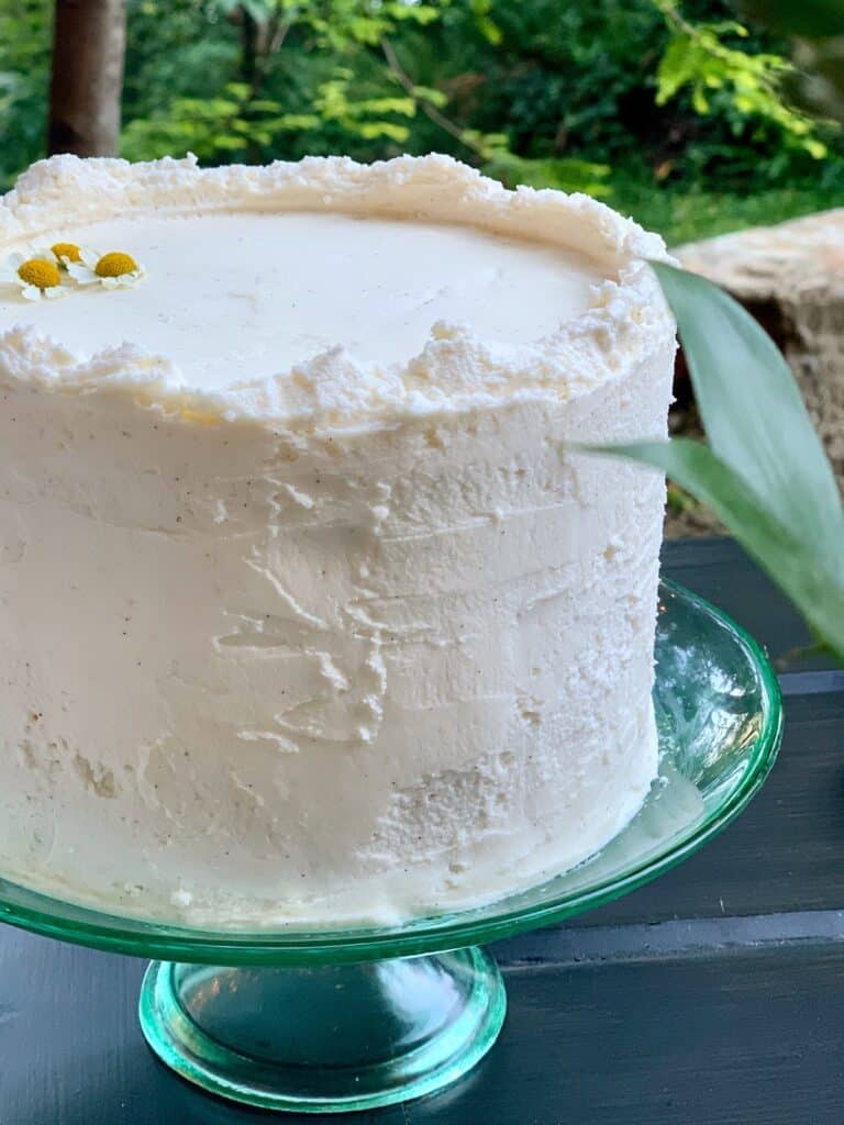 A layered carrot cake on a green glass pedestal is covered with Cream Cheese Frosting and topped with chamomile blossoms to make a beautiful wedding cake.
