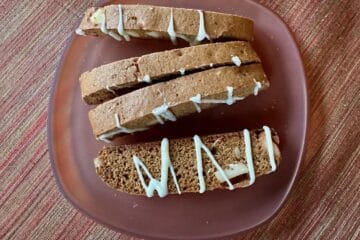 Gingerbread Biscotti drizzled with white chocolate are arranged with top and side featured on a small amber plate.