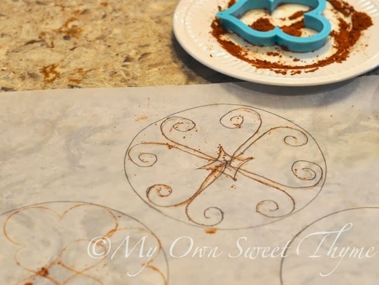 Filigree pattern for piped meringue layer designed with cookie cutters and cocoa powder on sheets of parchment paper. 